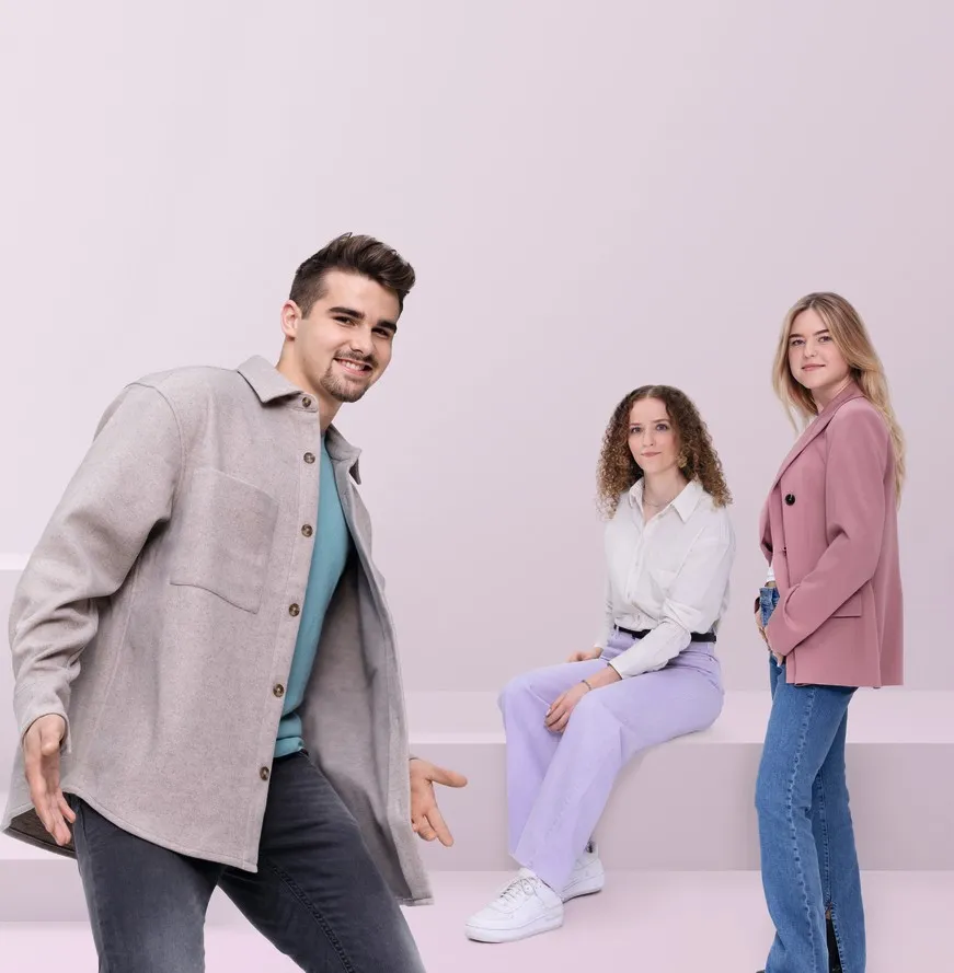 Drei junge Erwachsene lächeln in die Kamera, einer steht im Vordergrund, zwei sitzen und stehen im Hintergrund vor einem hellen Hintergrund.