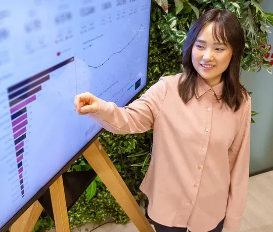 A smiling woman in a pink shirt points at a data chart on a screen, in a lively and green office space.