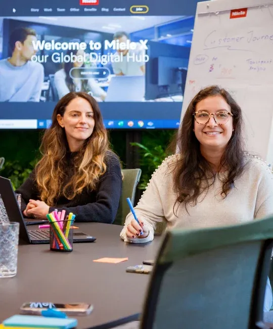 Two smiling women at a conference table with a laptop and colorful pens, ready for a creative session at Miele's Digital Hub.