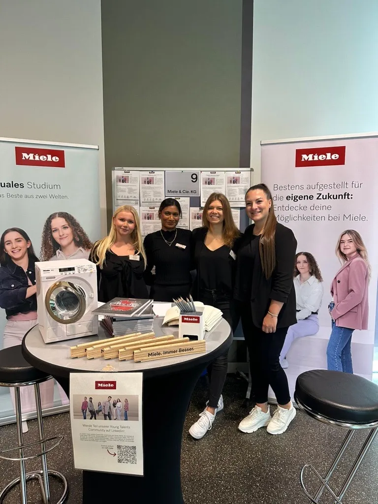 Vier junge Frauen stehen an einem Miele-Stand auf einer Karrieremesse, umgeben von Informationsmaterialien und Bannern.