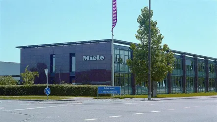 Front view of Miele's modern building in Denmark with glass facade and company sign, set against a clear blue sky.