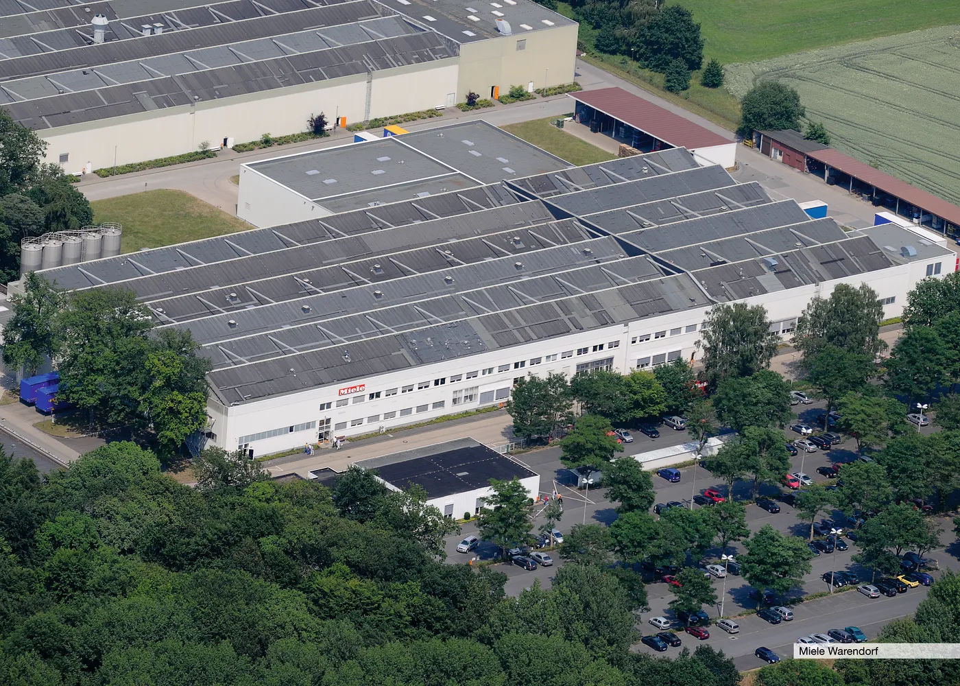 Luftaufnahme des Miele-Werksgebäudes, umgeben von Bäumen und Parkplätzen, ideal für eine Karriere im Bereich der Fertigung.