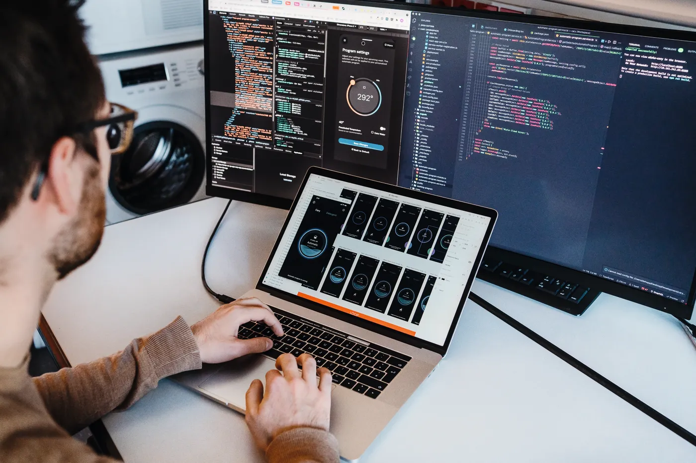 Ein Softwareentwickler arbeitet an einem Laptop und zwei Monitoren mit Code und Benutzeroberflächen-Designs, im Büro von Miele & Cie. KG.