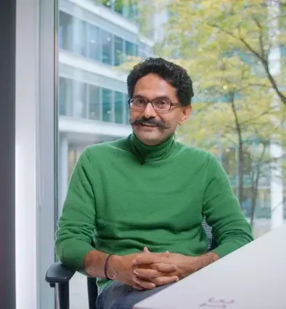 A friendly man in a green sweater sits at a desk by a large window in a modern office building.