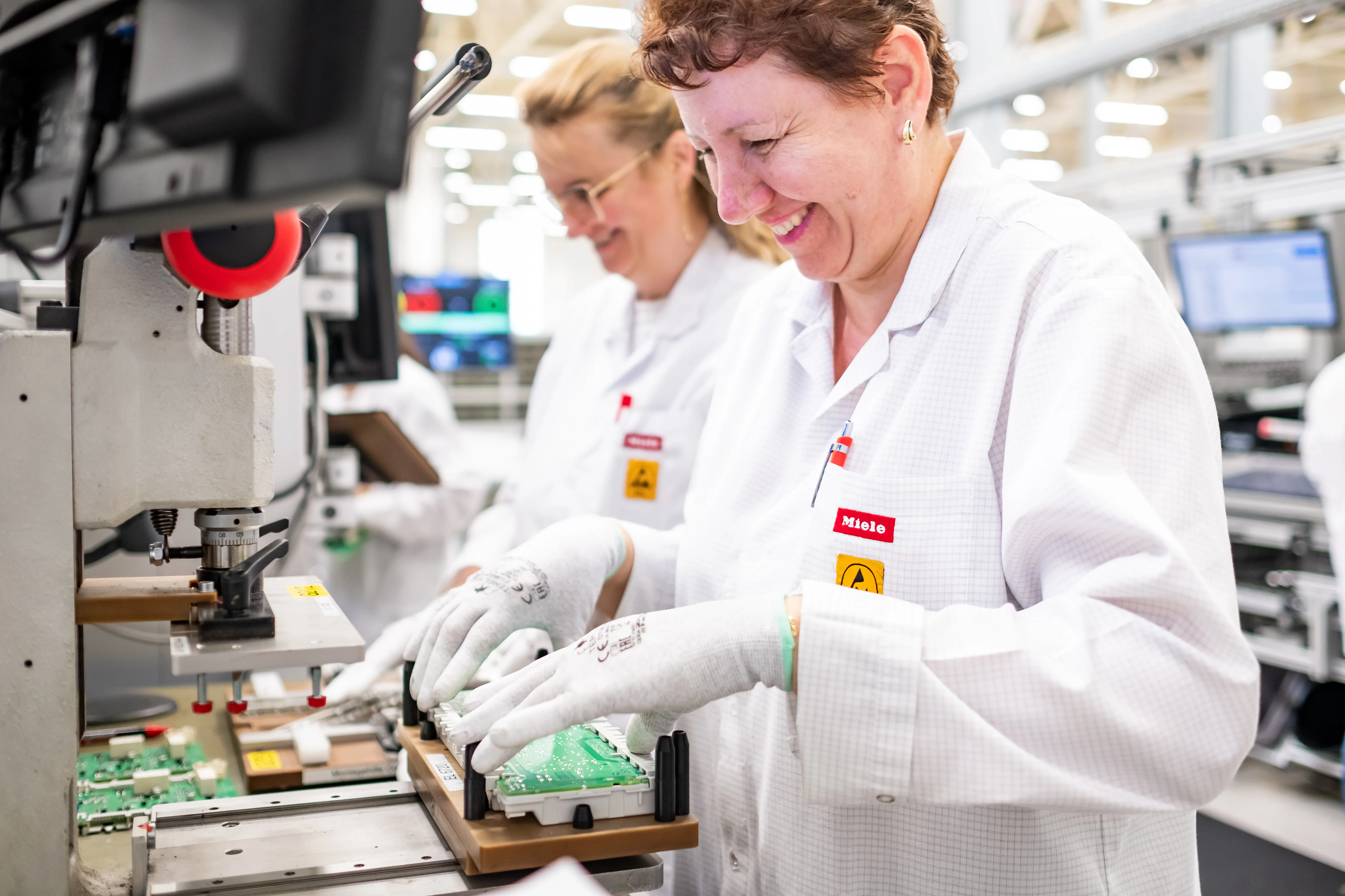 Lucrătoare fericite asamblează plăci electronice într-o fabrică Miele, în halate albe de laborator.
