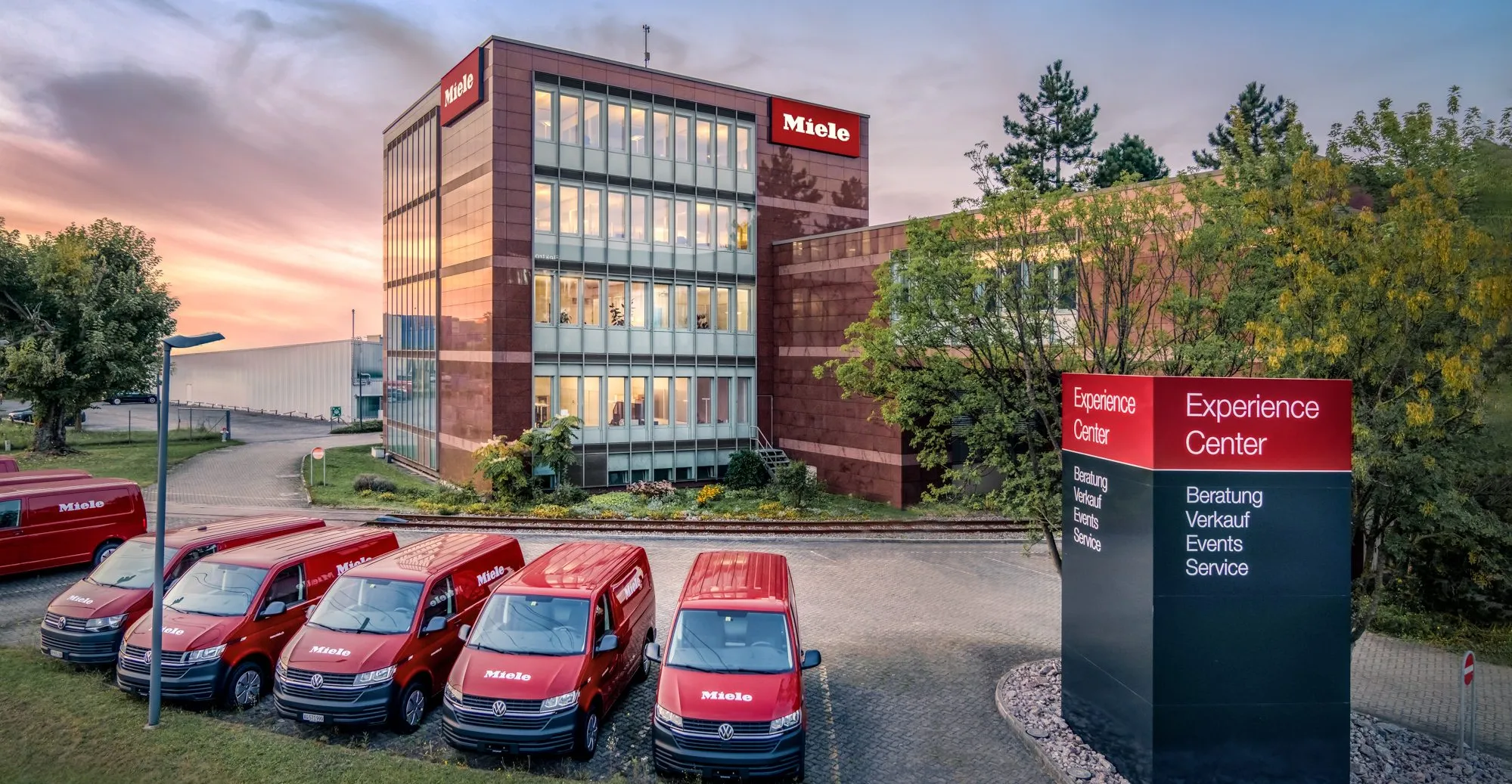 Gebäude der Miele & Cie. KG mit roten Lieferwagen und einem Schild für das Experience Center bei Sonnenuntergang.