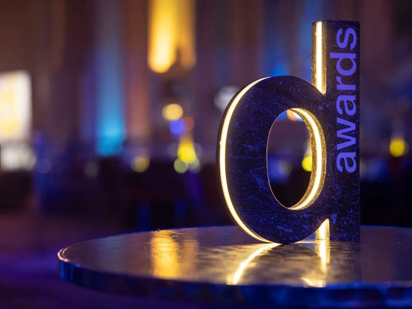 Close-up of a stylish illuminated 'd' award statue on a reflective table, set against a vibrant, colorful background.