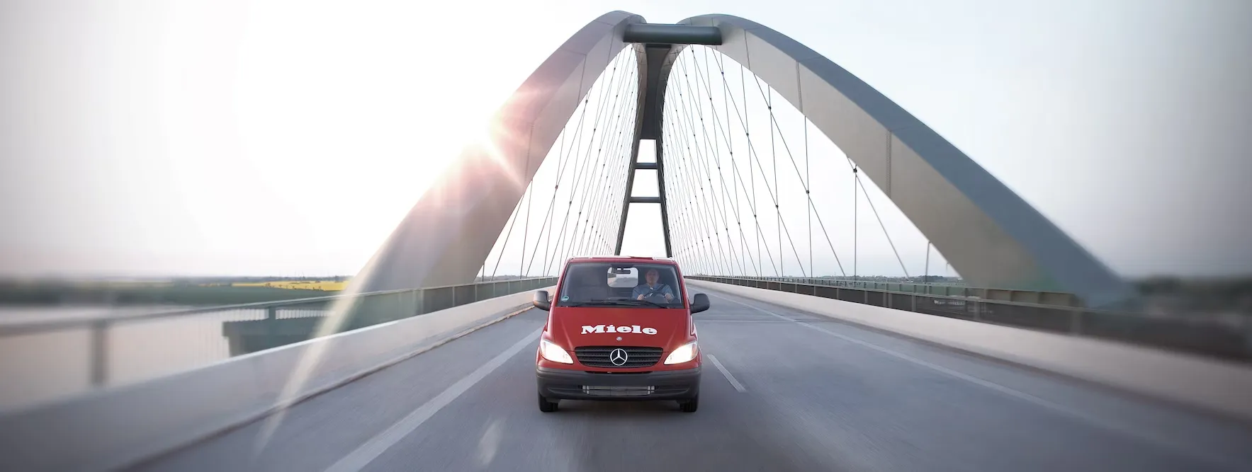 Red Miele van crossing a modern bridge under the sun, showcasing Miele's commitment to quality and innovation.