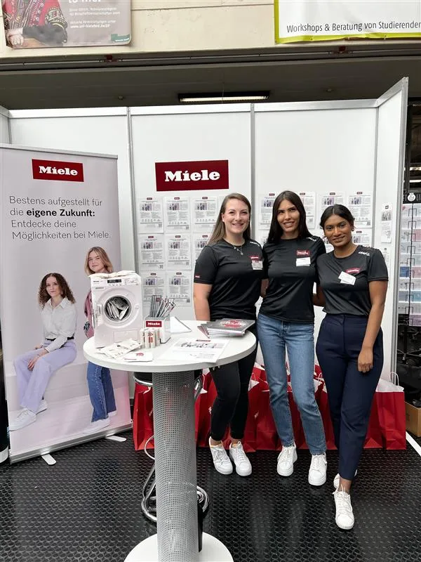 Drei Mitarbeiterinnen am Miele-Messestand lächeln. Der Stand zeigt Broschüren und Werbematerialien. Ein Plakat bewirbt Karrierechancen.