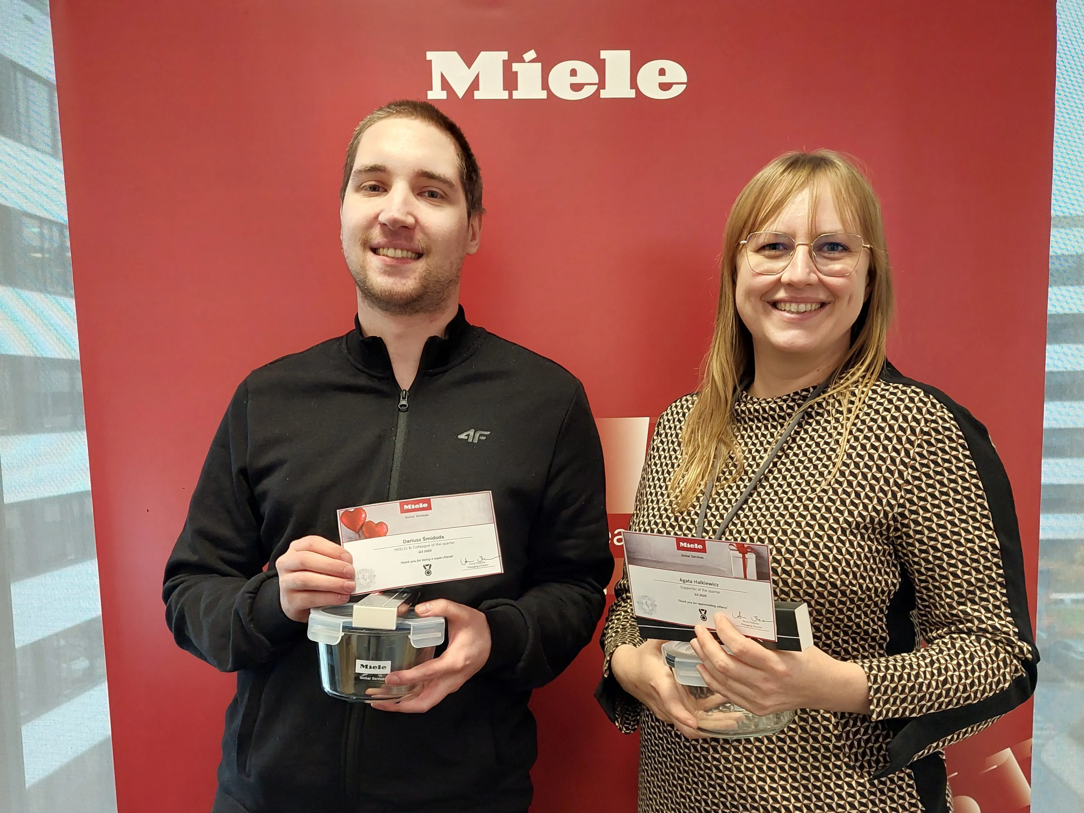 Two Miele employees smiling and holding awards in front of a Miele backdrop, celebrating achievements in manufacturing.