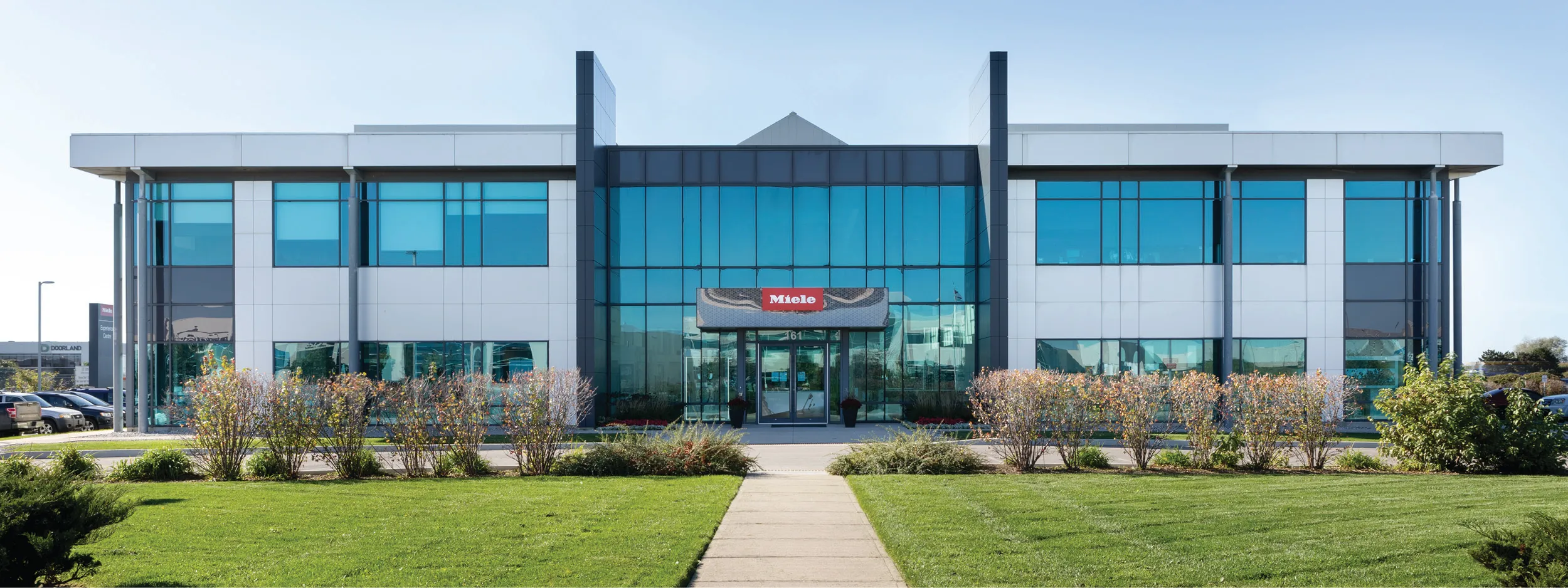 Front view of Miele's sleek, modern office building in Canada with glass facade, surrounded by lush greenery.