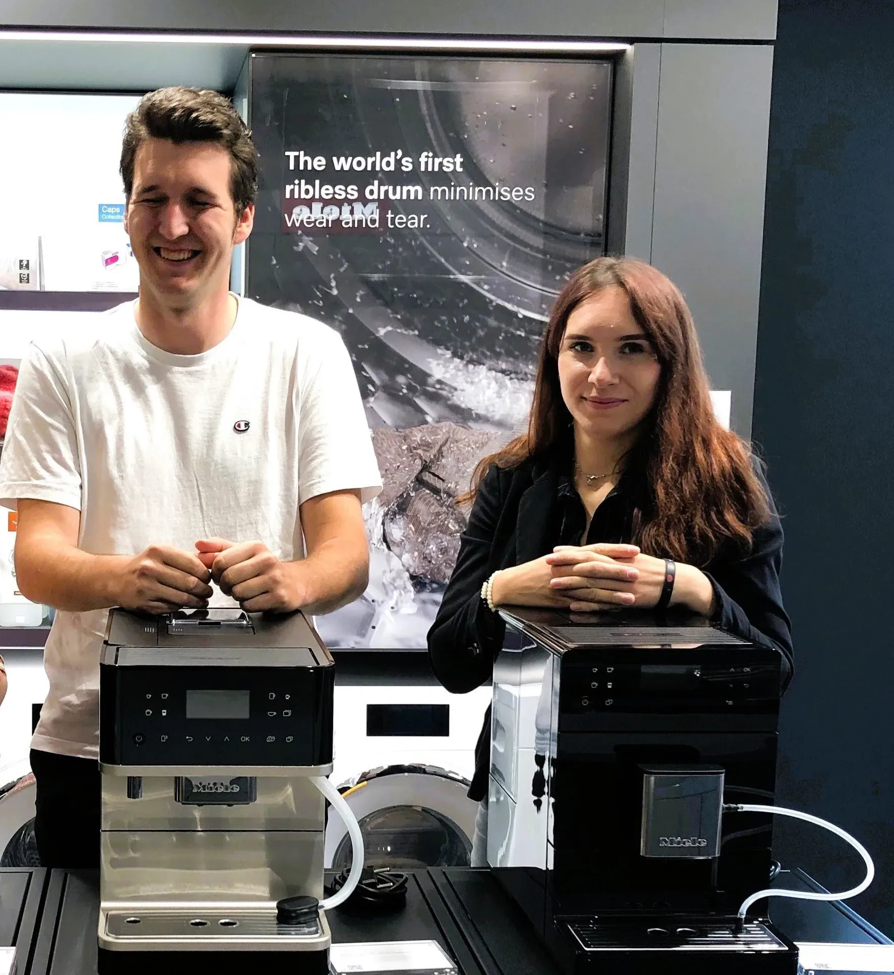 Two young professionals smiling beside Miele coffee machines, showcasing innovation at a manufacturing event.