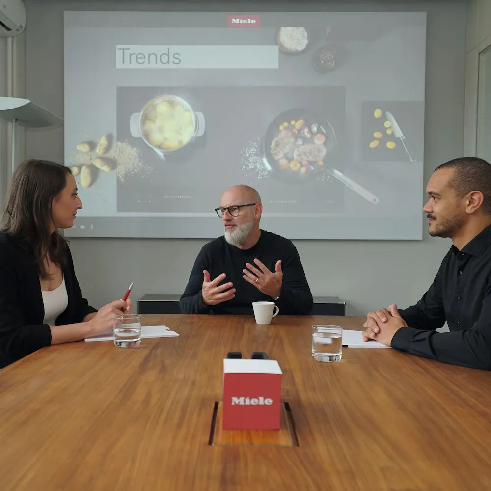 Drei Personen sitzen an einem Konferenztisch und sprechen miteinander, im Hintergrund ist eine Präsentation auf einer Leinwand zu sehen