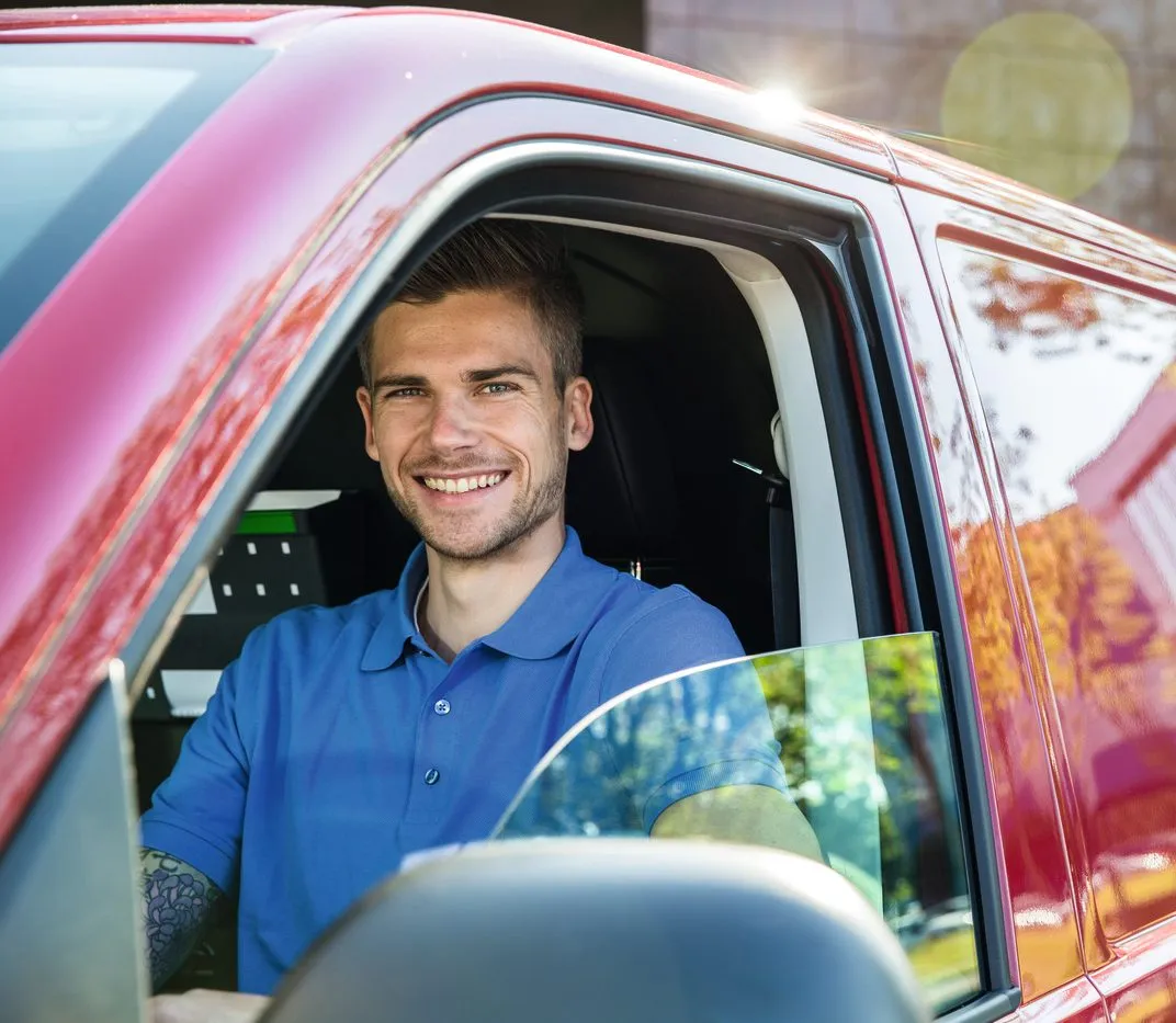 A young man in a blue polo shirt, smiling while sitting in a red vehicle, ready for his next delivery.