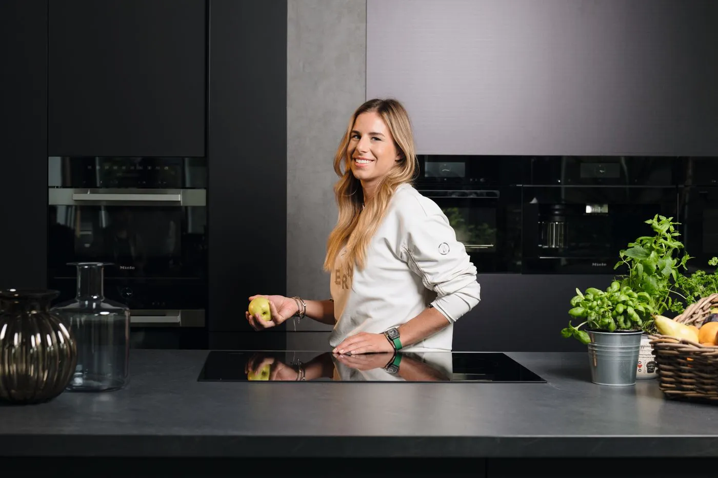A woman smiling in a modern kitchen with Miele appliances, holding an apple and surrounded by fresh herbs and fruit.