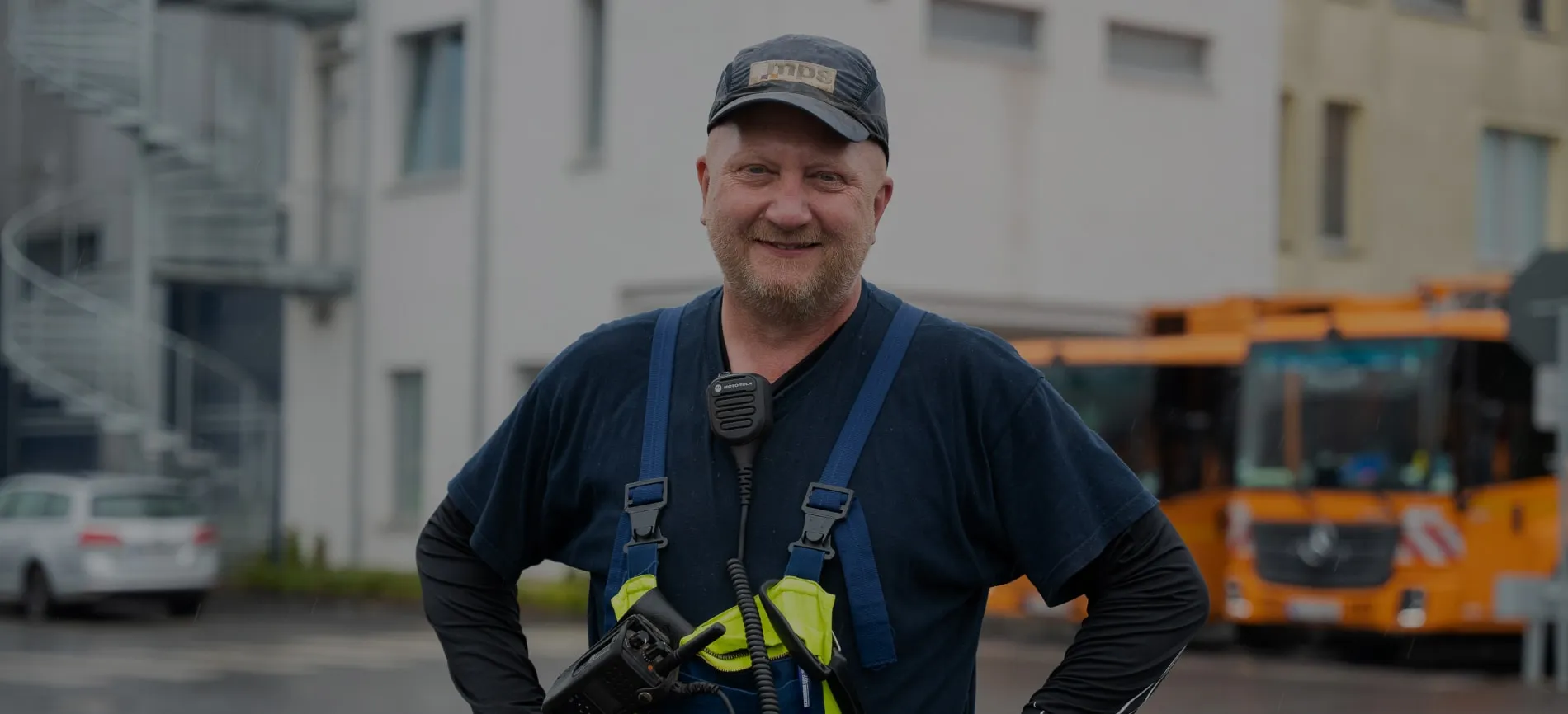 Ein lächelnder Bauarbeiter in Arbeitskleidung und MPS-Kappe steht auf dem Firmengelände vor Bau- und Einsatzfahrzeugen.