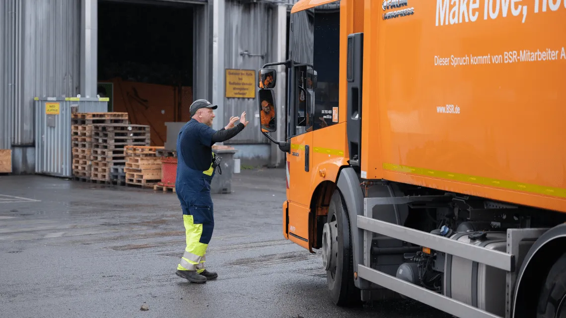 Ein Kollege weist einen Fahrer in einem orangefarbenen Lkw auf dem Betriebsgelände ein.