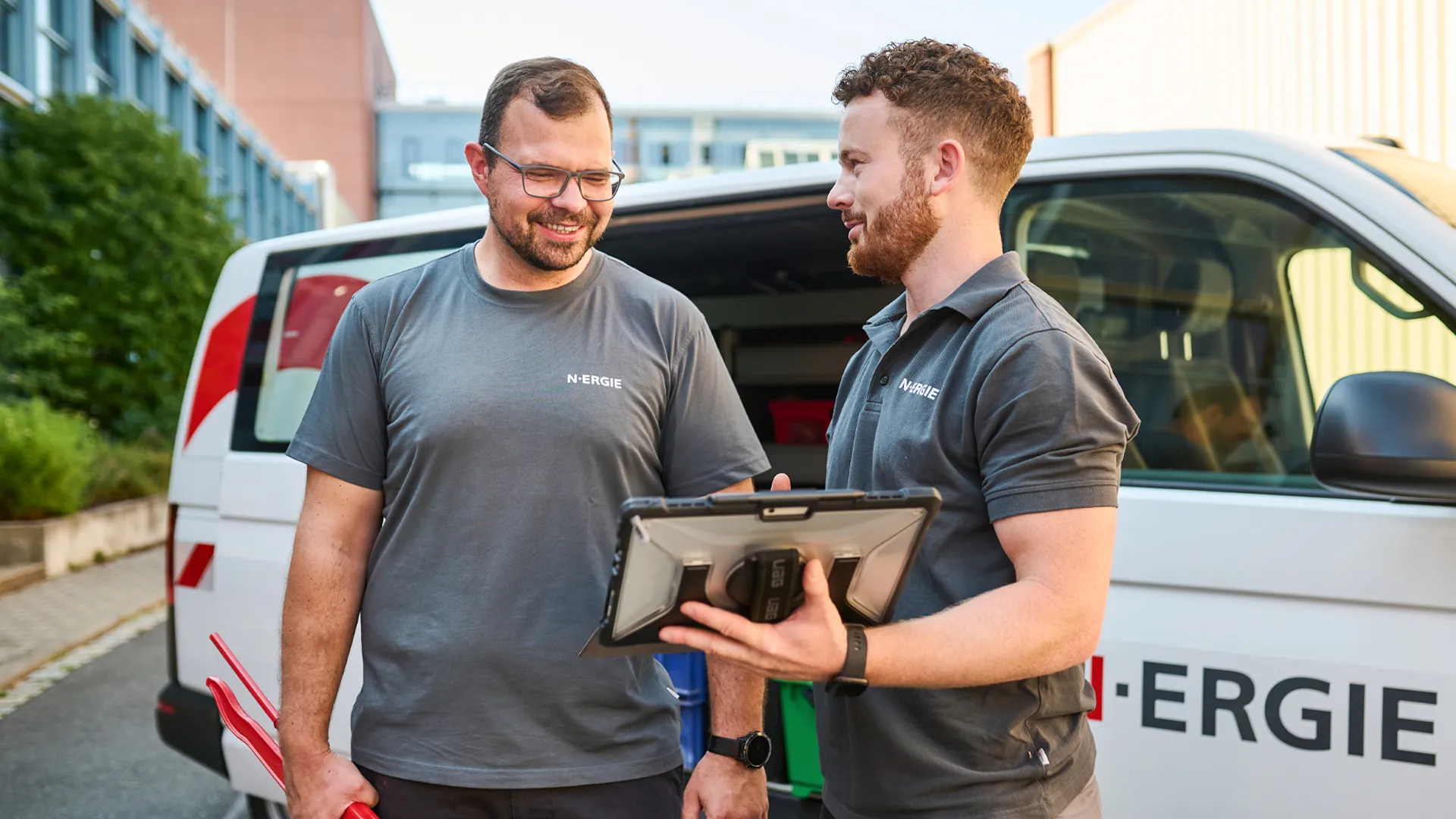 Zwei N-ERGIE-Mitarbeiter im Gespräch vor einem Firmenfahrzeug, einer hält ein Tablet. Perfekt für deine Zukunft in der Energiebranche.