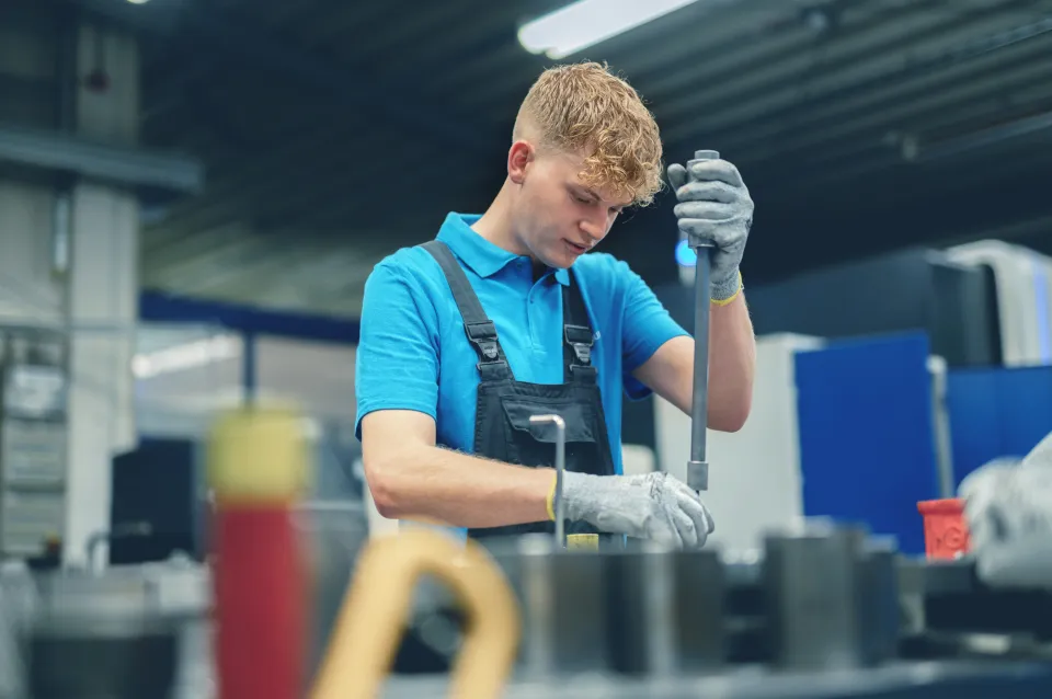 Ein junger Mann in Arbeitskleidung arbeitet konzentriert an einer Maschine in einer Fabrikhalle.
