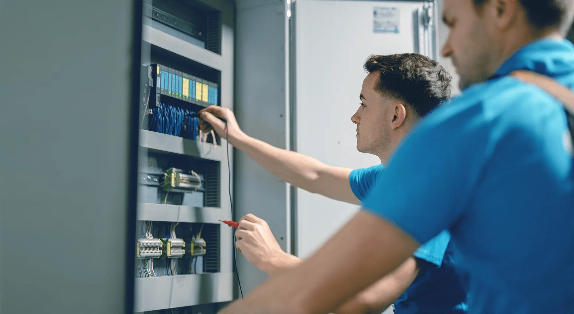 Zwei Techniker arbeiten an einem Schaltschrank mit Kabeln und Geräten in einer industriellen Umgebung.