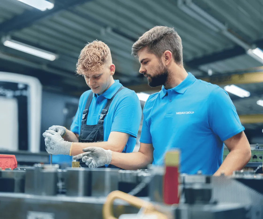 Zwei Arbeiter in blauen Poloshirts und Handschuhen arbeiten konzentriert an einer Maschine in einer Fabrikhalle.