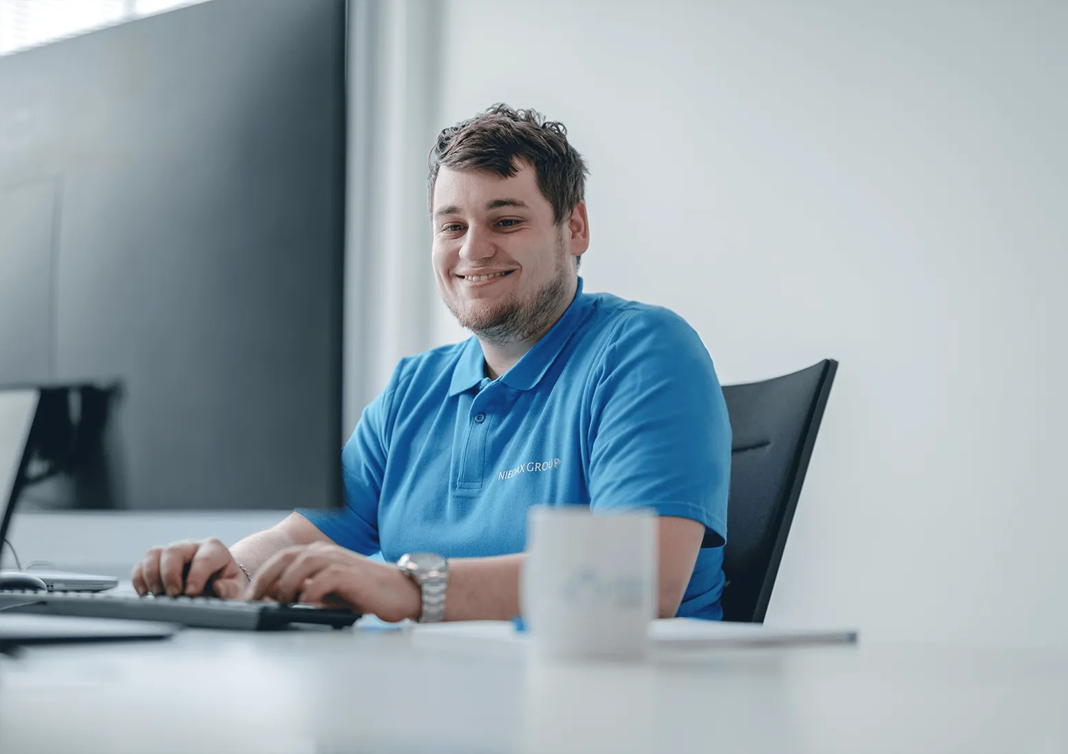 Ein lächelnder Mann in einem blauen Polohemd arbeitet am Computer in einem modernen Büro.