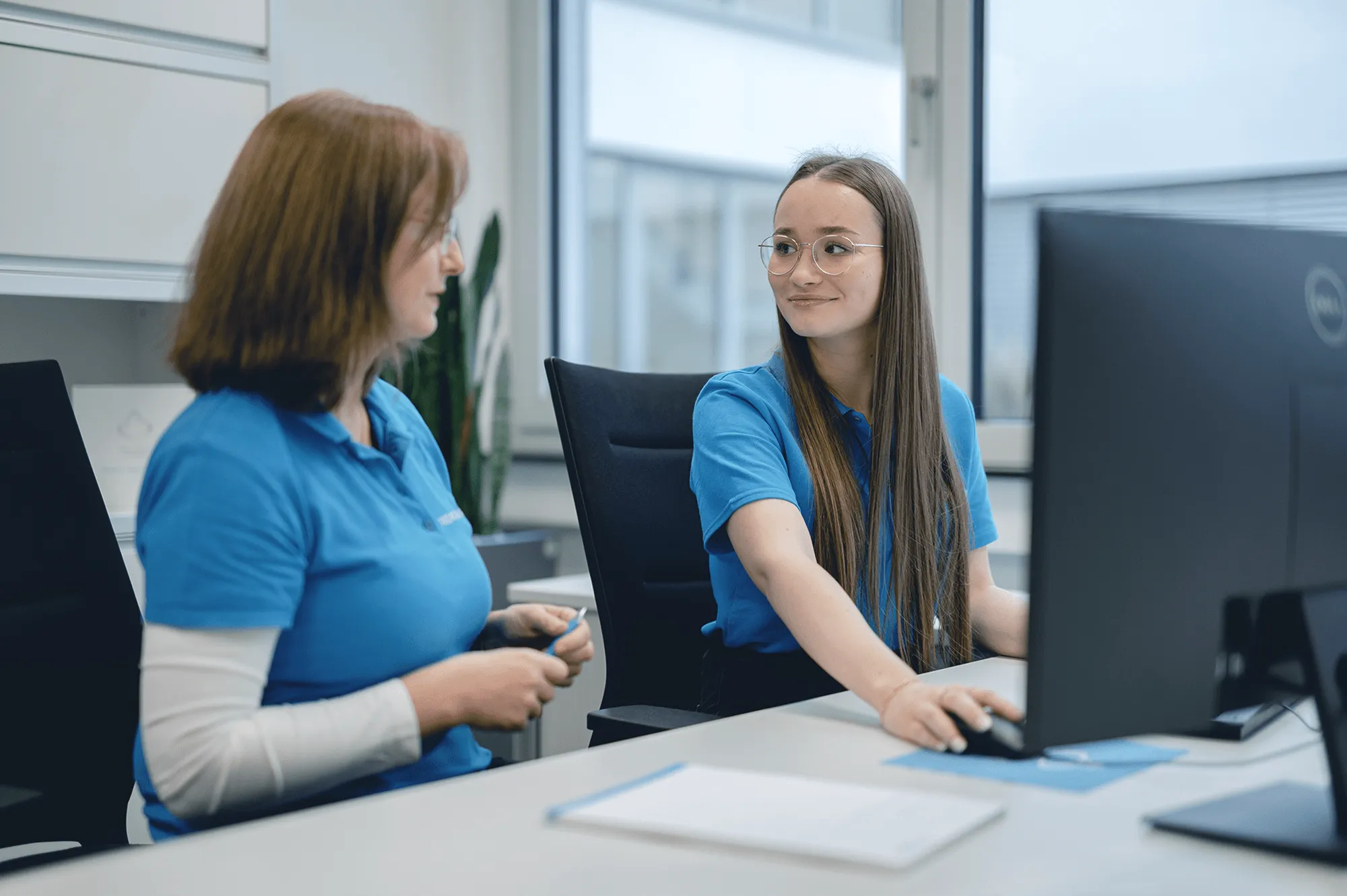 Zwei Mitarbeiterinnen in blauen Polohemden arbeiten gemeinsam am Computer in einem modernen Büro.