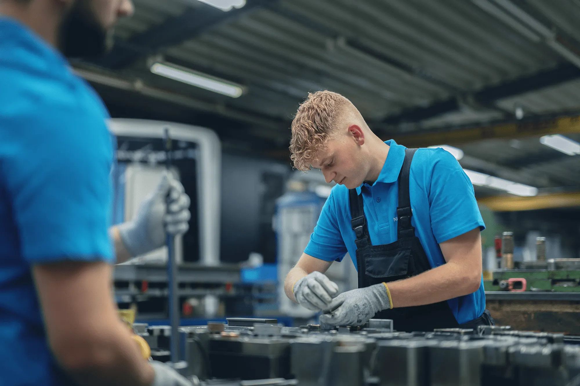 Ein junger Mann arbeitet konzentriert in einer Fabrikhalle, trägt blaue Arbeitskleidung und Handschuhe.