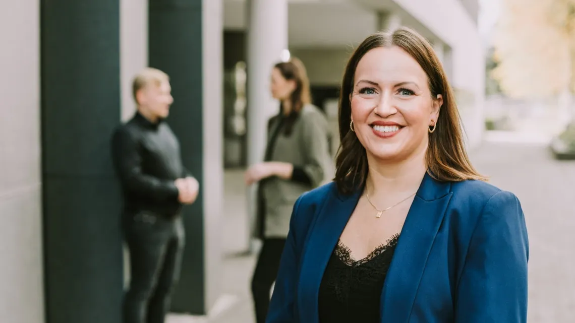 Junge Frau im Business-Outfit lächelt freundlich vor einem modernen Bürogebäude, zwei Kollegen im Hintergrund im Gespräch.