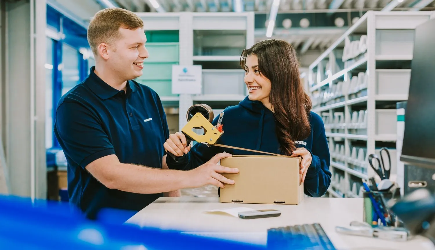 Zwei Mitarbeiter von NOWEDA verpacken gemeinsam eine Schachtel in einem Lager. Sie lächeln sich an.