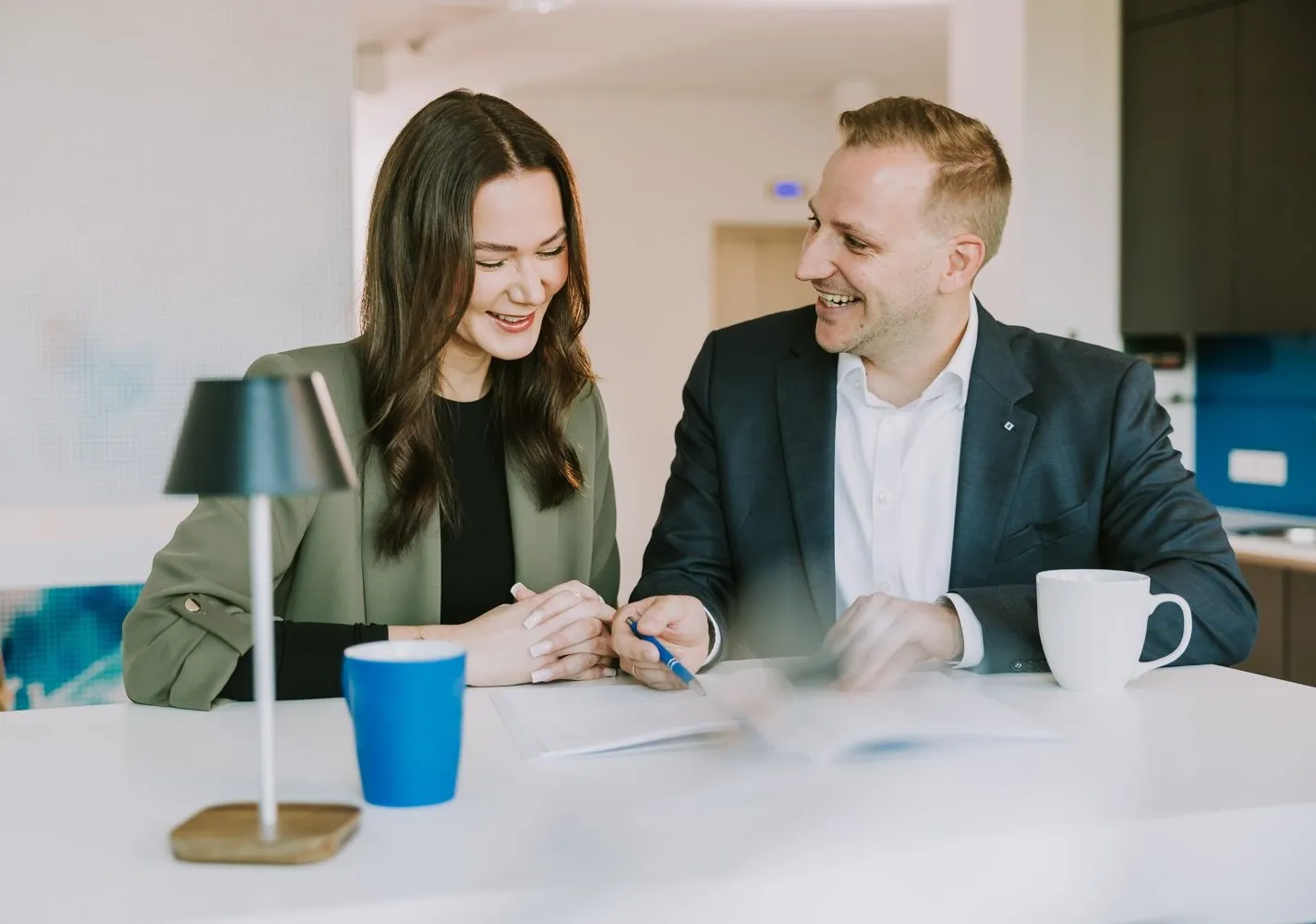 Ein Mann und eine Frau lachen und besprechen Dokumente bei einer Tasse Kaffee in einem modernen Büro.