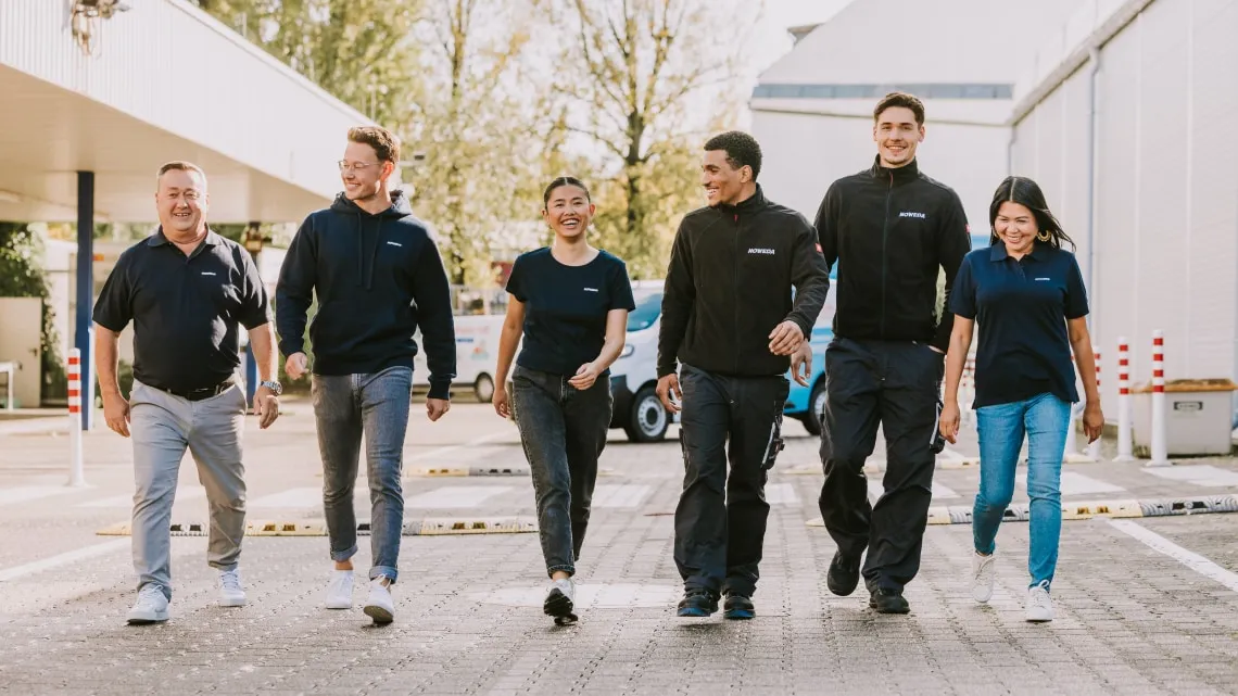 Sechs NOWEDA Mitarbeitende gehen gemeinsam lachend draußen, Teamgeist und Freude am Arbeitsplatz stehen im Fokus.