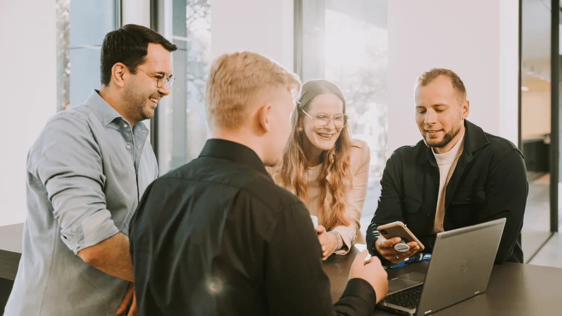 Vier junge Kolleg:innen lachen gemeinsam bei einem lockeren Meeting am Tisch im NOWEDA Büro, Laptop und Smartphone dabei.