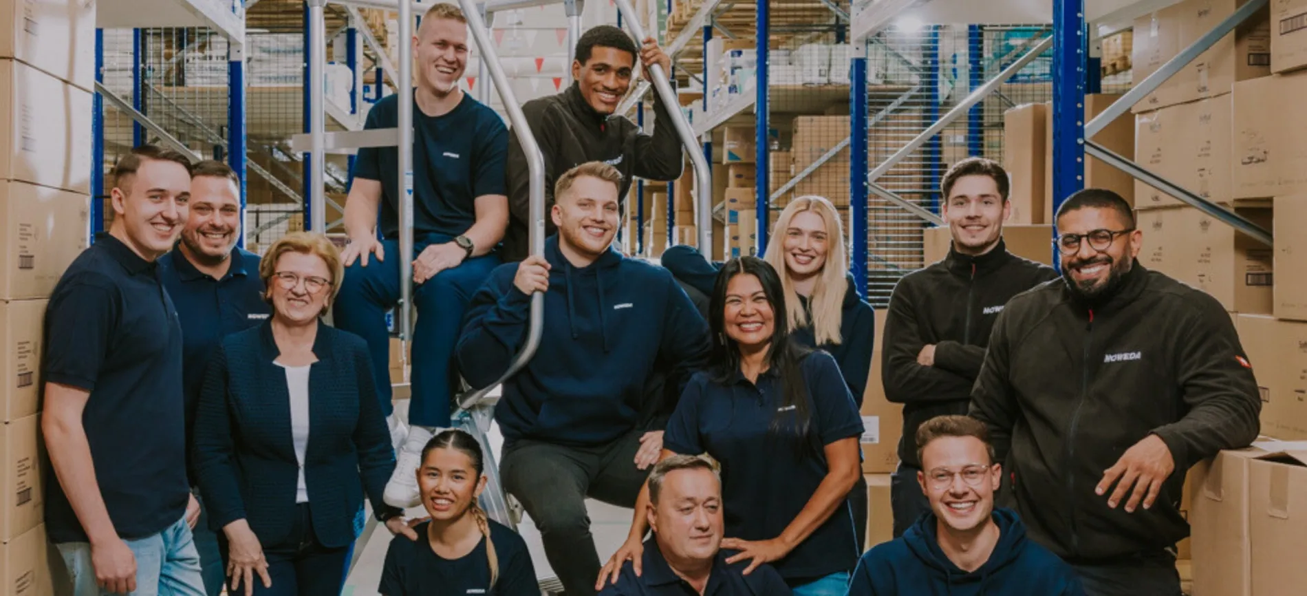 Ein fröhliches NOWEDA-Team posiert gemeinsam im Lager zwischen Kartons und Regalen, bereit für neue Kollegen wie dich.