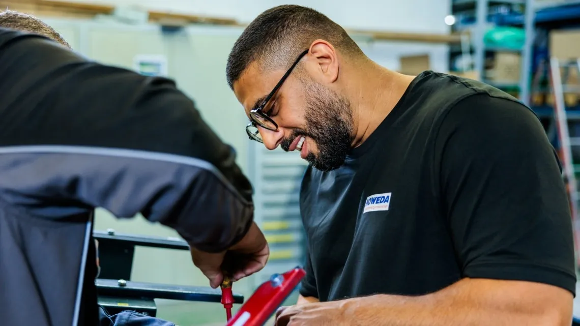 Ein Mann im NOWEDA-Shirt arbeitet konzentriert mit einem Kollegen an einer technischen Aufgabe in einer Werkstatt.