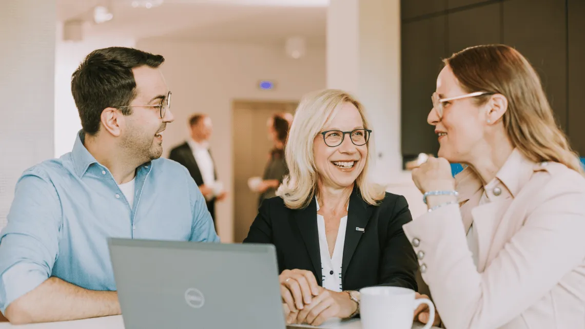 Drei NOWEDA-Mitarbeitende lachen und tauschen sich an einem Tisch im Büro aus – Teamarbeit im Pharma-Bereich.