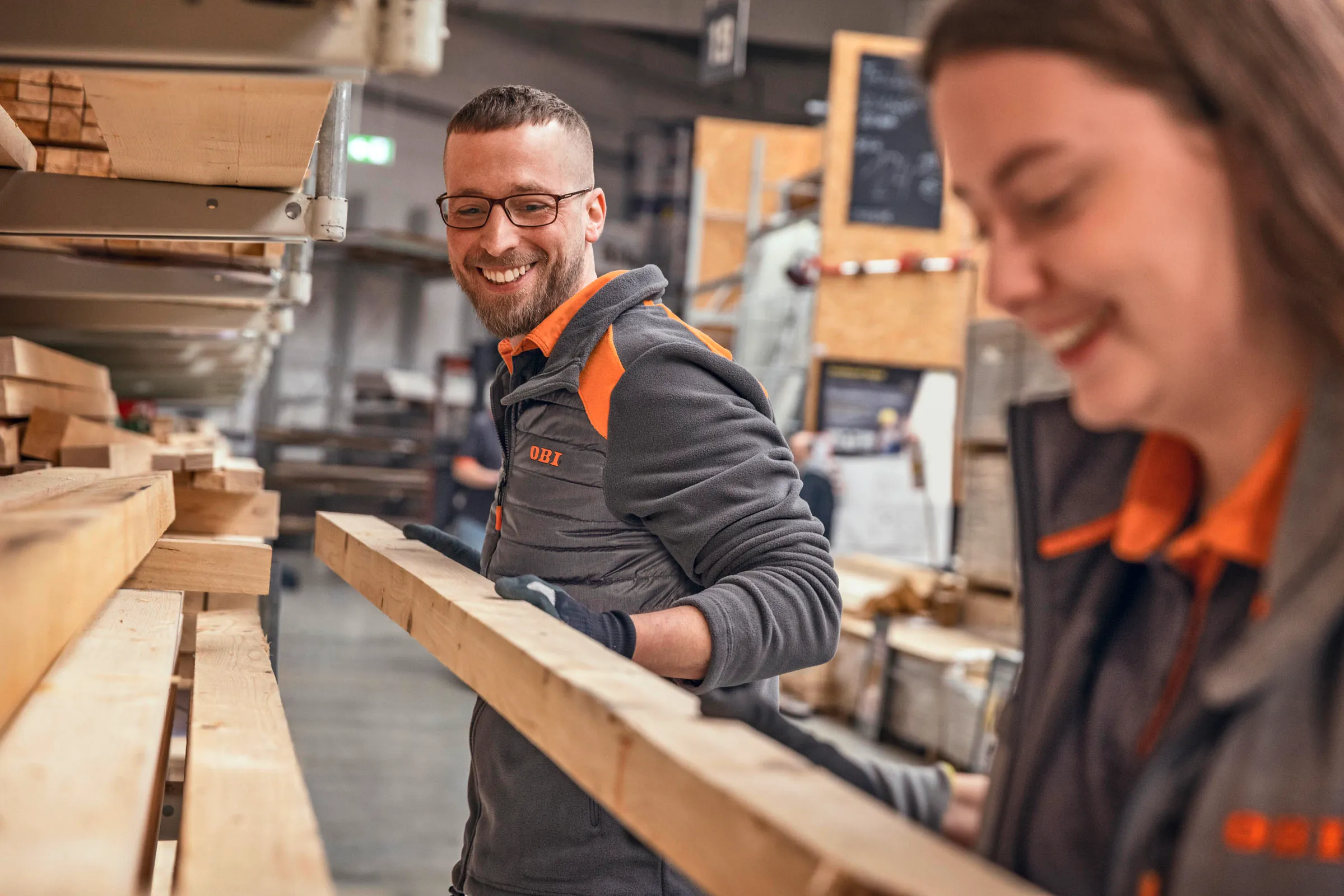 Zwei OBI-Mitarbeitende sortieren gemeinsam Holzbretter im Lager und haben Spaß bei der Arbeit.