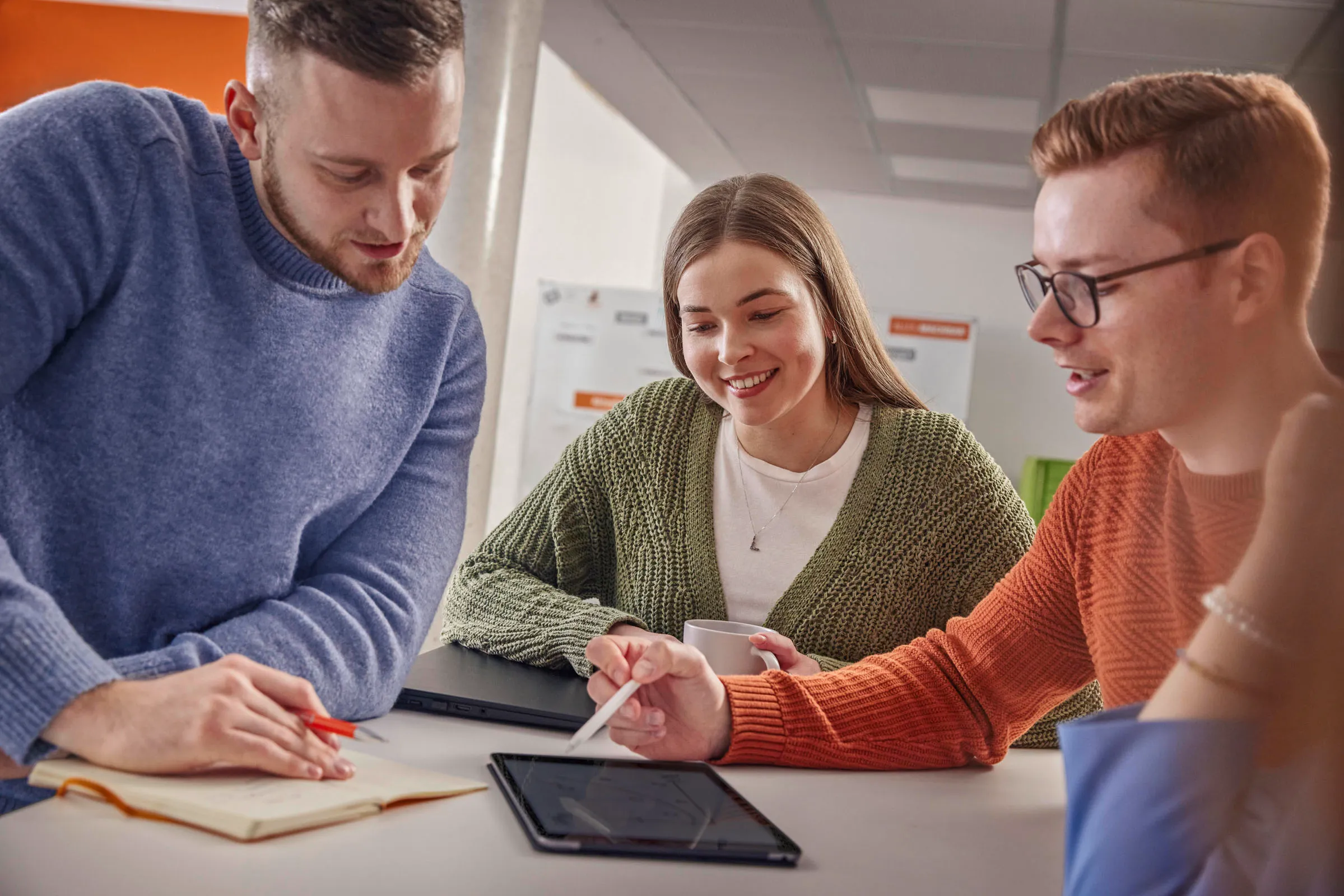 Drei junge Menschen arbeiten gemeinsam an einem Tablet und Notizbuch im modernen OBI Büro, lächeln und tauschen Ideen aus.
