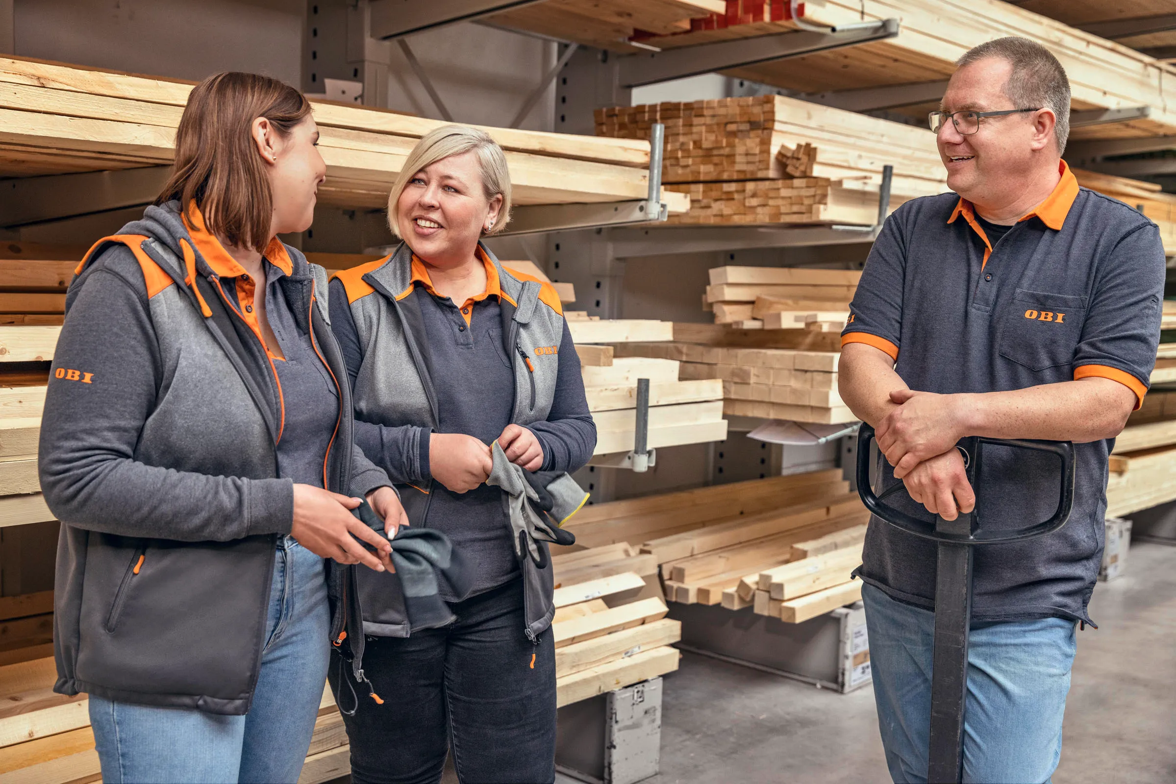 Drei OBI-Mitarbeitende unterhalten sich freundlich im Holzlager eines Baumarkts.