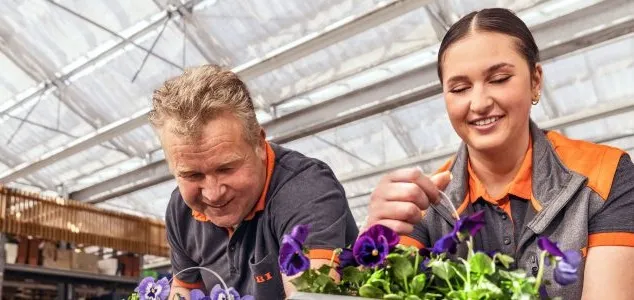 Zwei OBI-Mitarbeiter pflanzen gemeinsam Blumen in einem Gewächshaus.