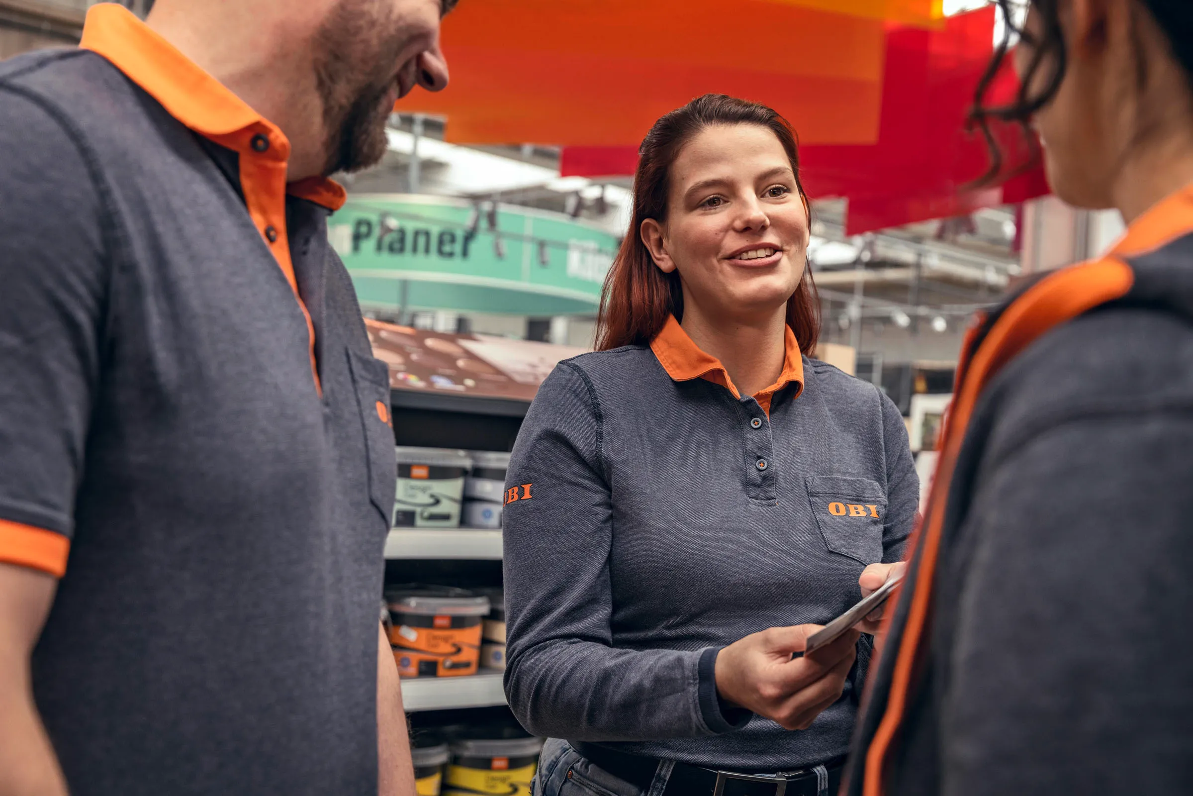Drei OBI-Mitarbeiter im Gespräch im Baumarkt, eine Kollegin lächelt und informiert Dich freundlich.