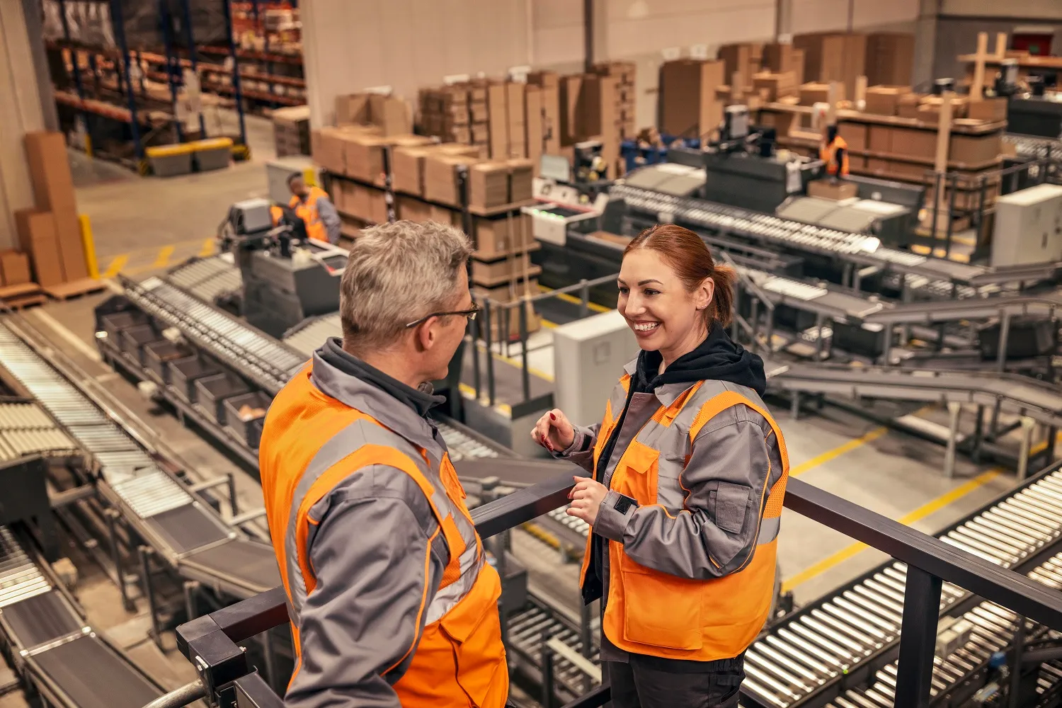Zwei Mitarbeitende im OBI-Logistikzentrum unterhalten sich freundlich in Arbeitskleidung vor Förderbändern.