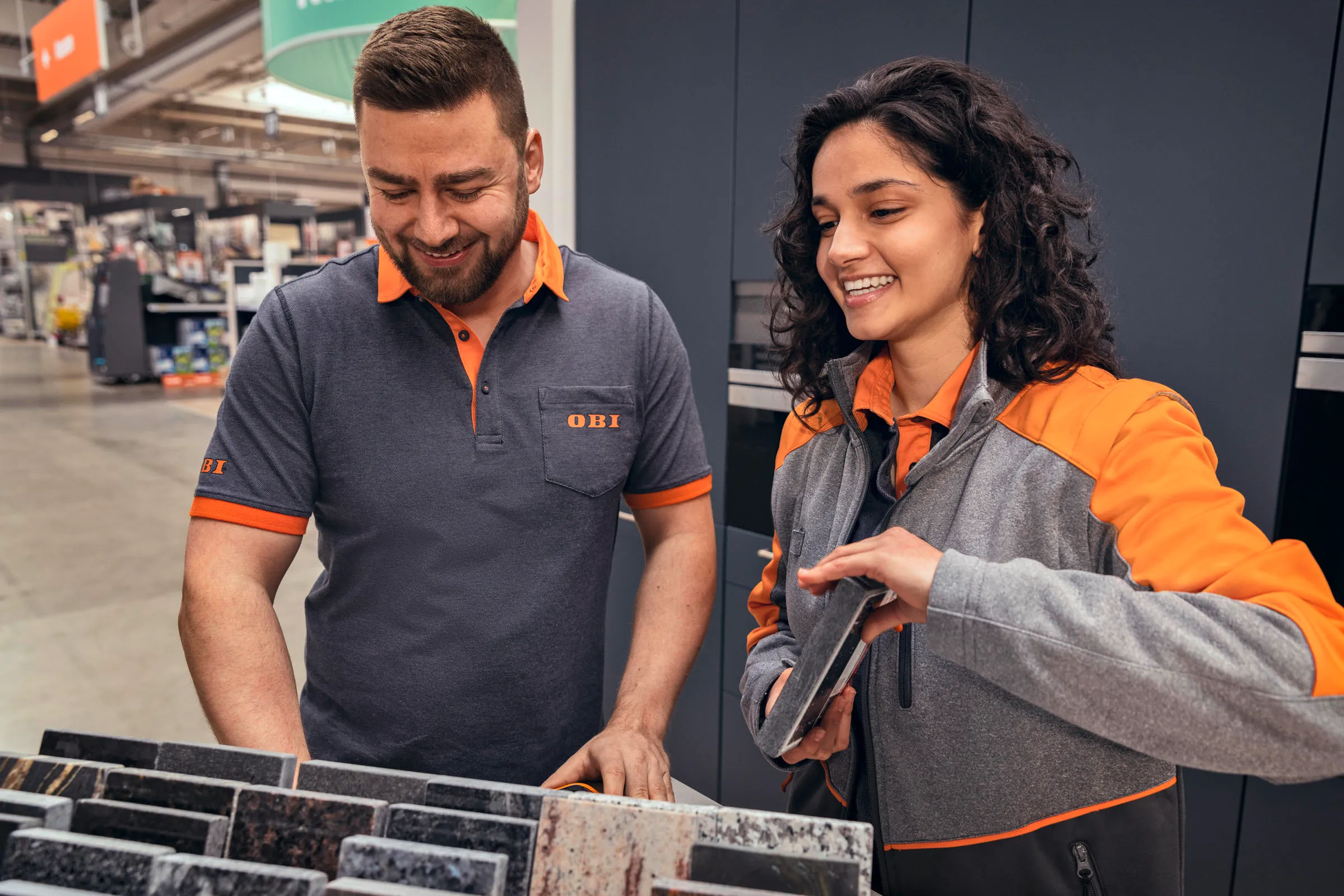 Zwei OBI-Mitarbeitende beraten sich lächelnd bei der Auswahl von Fliesenmustern im Baumarkt.