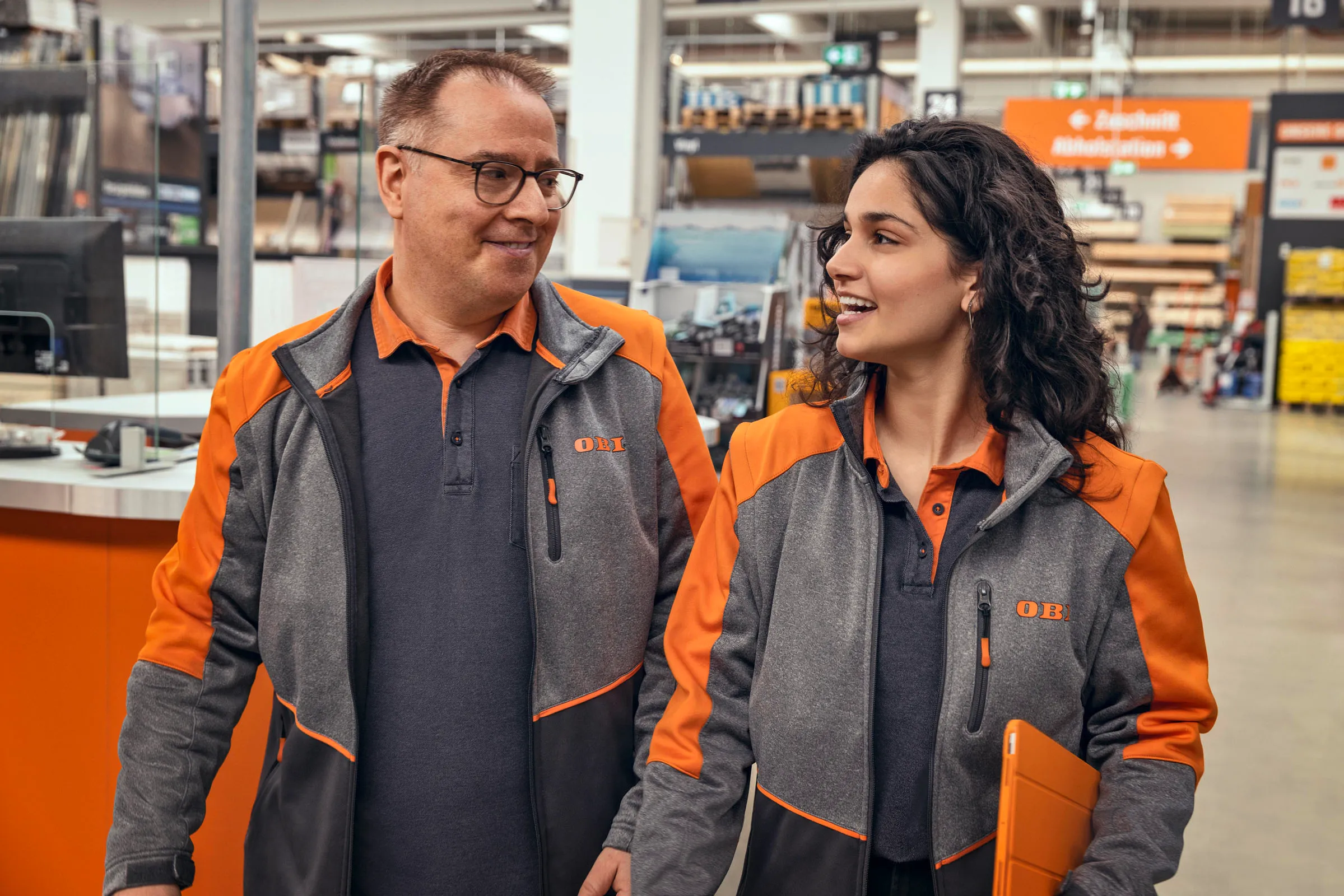 Zwei OBI Mitarbeitende im Gespräch, lächelnd im Baumarkt, zeigen Teamarbeit und freundliche Arbeitsatmosphäre.