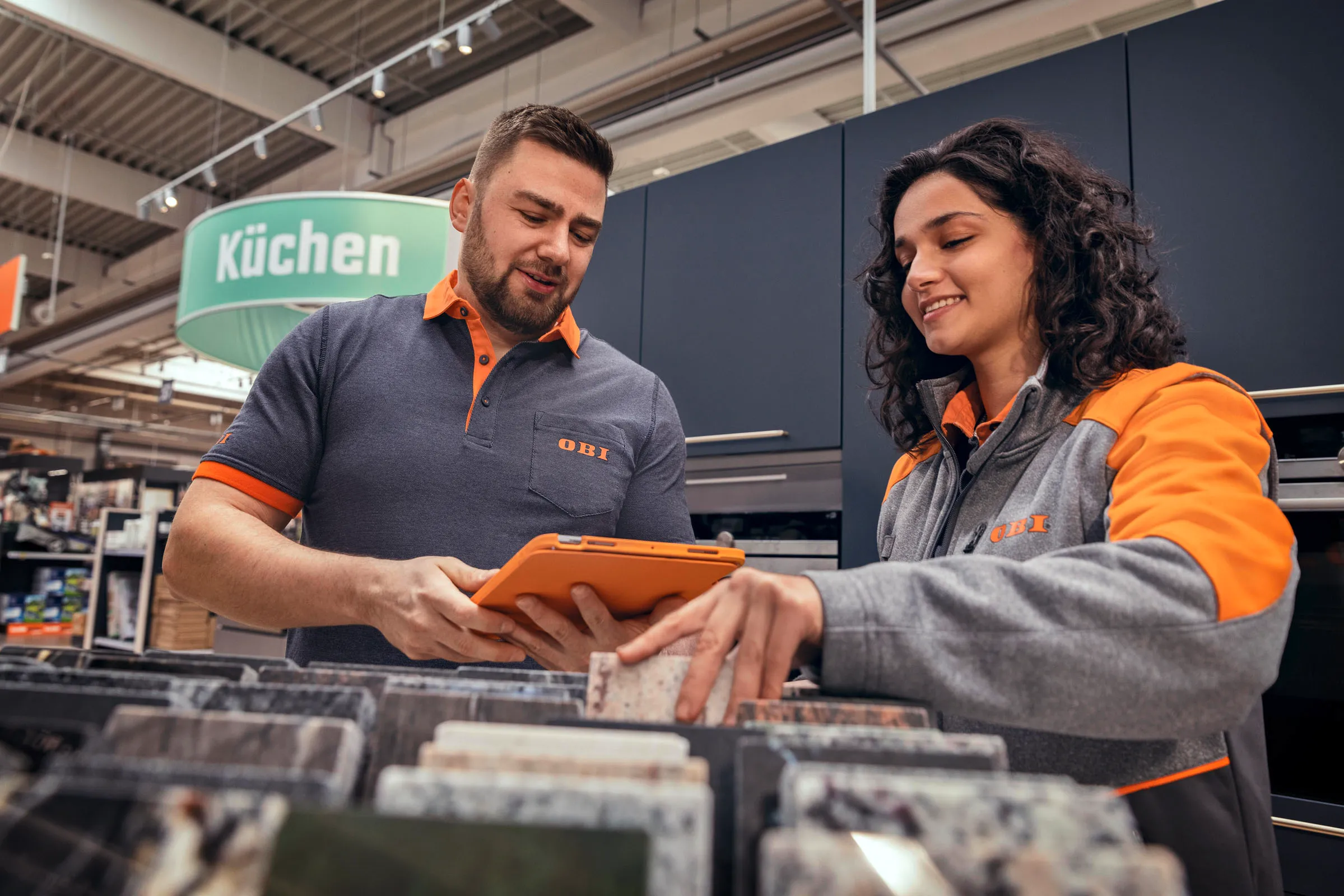 Zwei OBI-Mitarbeiter beraten Dich freundlich bei der Auswahl von Stoffmustern in einem modernen Baumarkt.