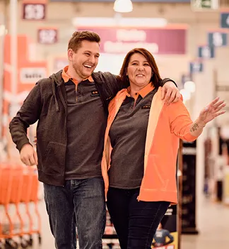 Zwei fröhliche Mitarbeiter in OBI-Uniform gehen durch einen Baumarkt.