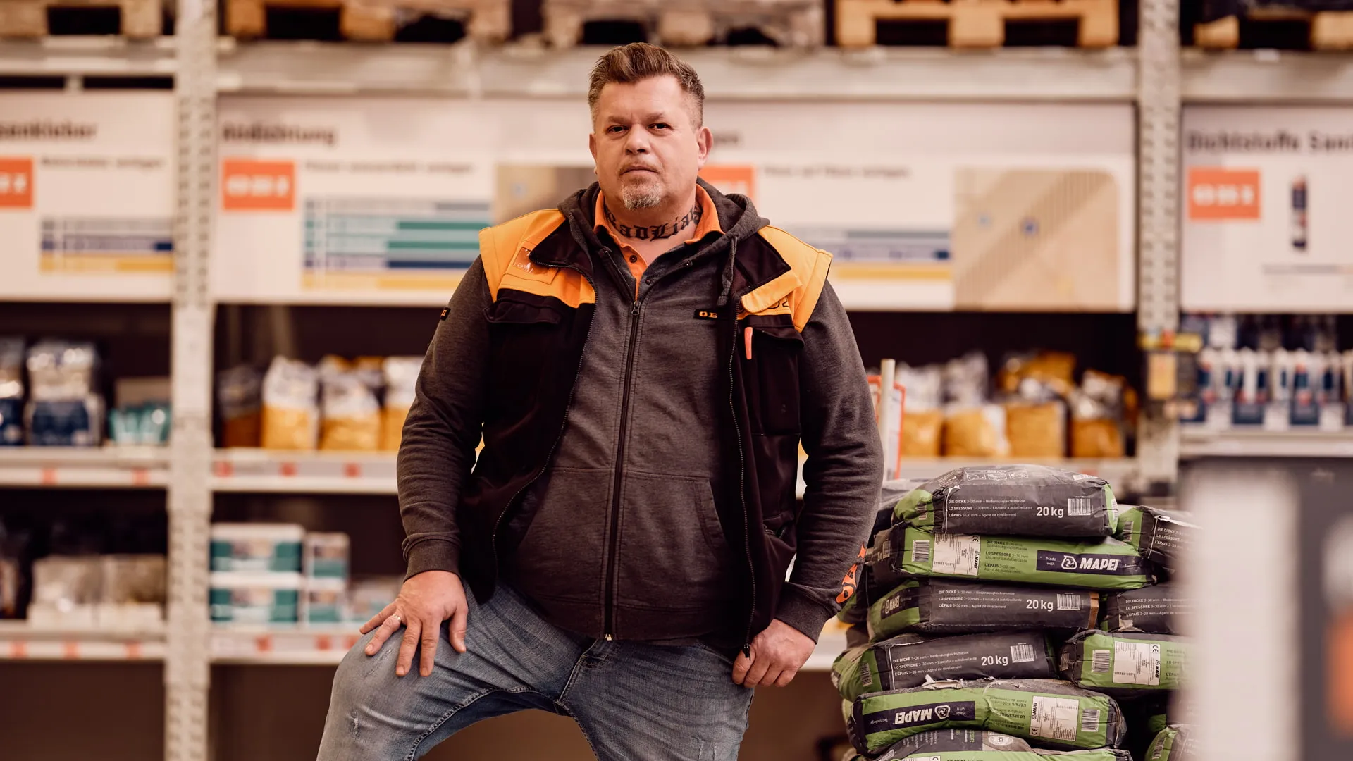 Ein OBI Mitarbeiter steht im Baumarkt vor Regalen mit Baustoffen und spricht Dich als zukünftige Kollegin oder Kollegen an.
