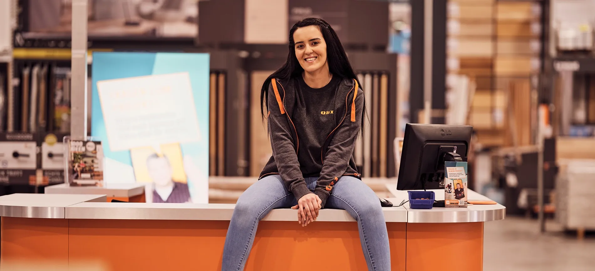 Eine lächelnde Mitarbeiterin von OBI AT GmbH sitzt entspannt auf einem Tresen in einem Baumarkt.