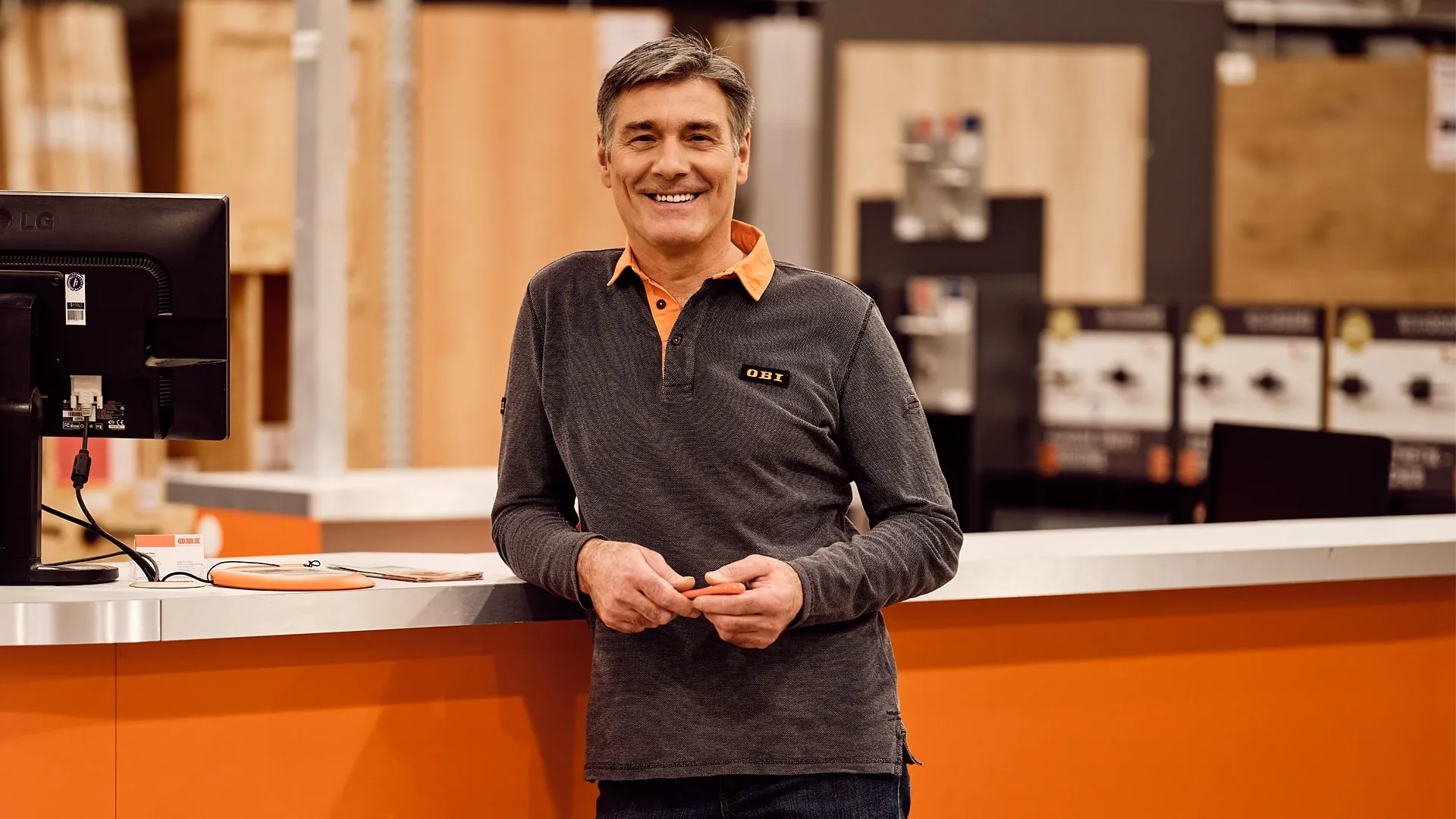 Ein Mitarbeiter von OBI AT GmbH lächelt an einem Verkaufstresen, umgeben von Holz und Baumaterialien.