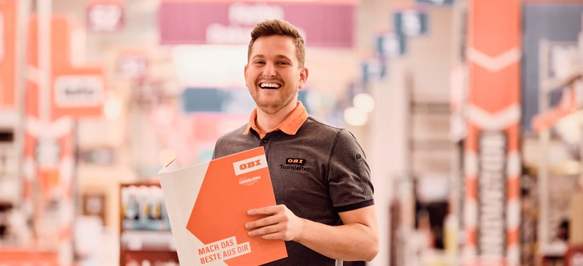 Lächelnder OBI Teamleiter im Markt mit Broschüre „Mach das Beste aus dir“ – Karriere bei OBI AT GmbH.